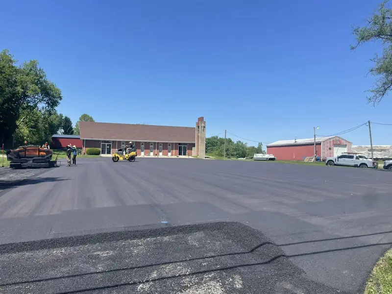 New asphalt layer going down across a church parking lot, with the paver and roller working in the background