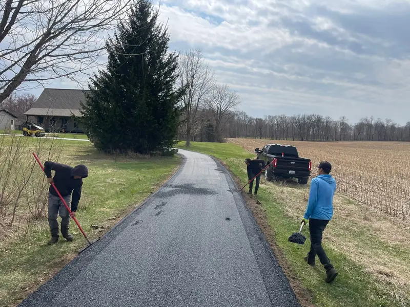 Crew raking and shaping the edges of a new driveway beside a harvested cornfield