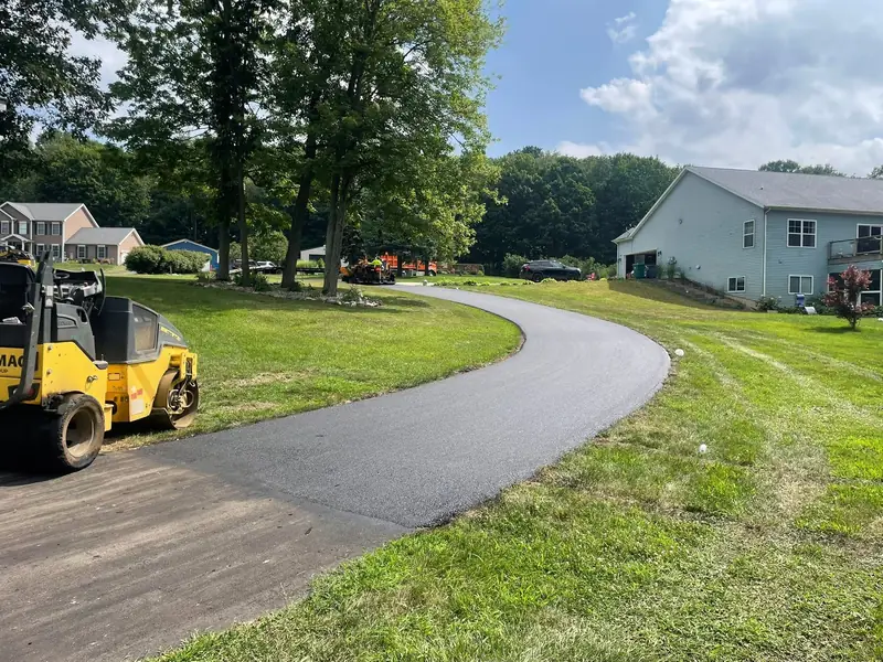 Tandem roller beside a new asphalt driveway that curves across the lawn toward a two-story home
