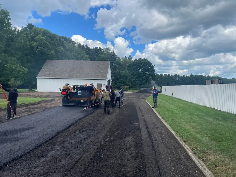 Crew raking hot mix behind the paver on a new driveway leading to a white garage
