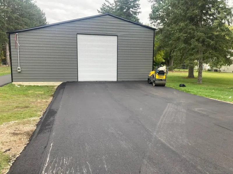 New asphalt apron in front of a gray steel garage, with the roller parked at the edge