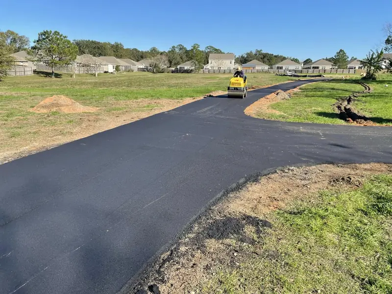 Roller finishing a long new asphalt drive across an open lot toward a row of homes