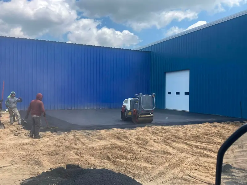 Crew finishing a new asphalt pad beside a blue steel building, with the roller on the fresh mat