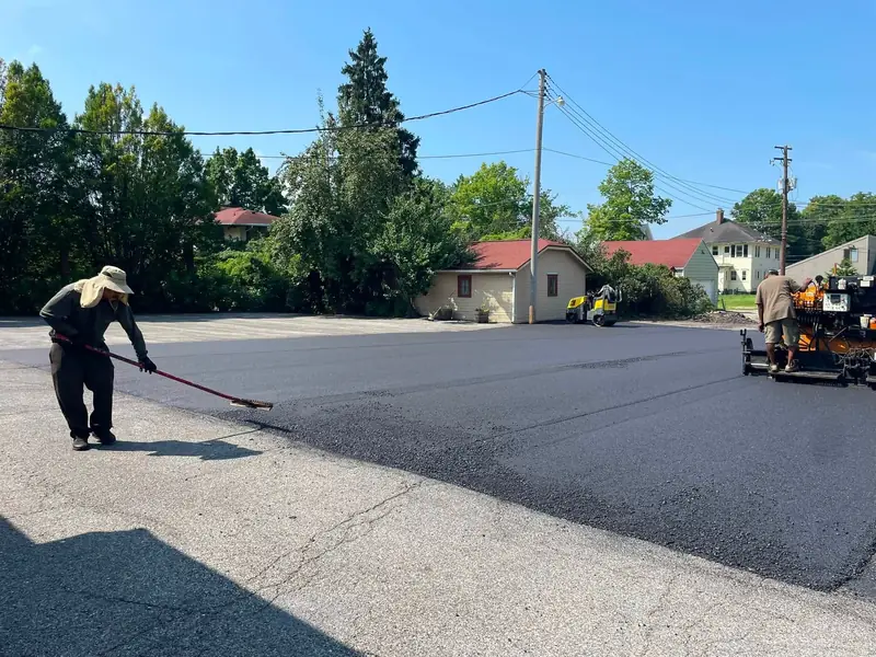 Crew member raking the edge of a new asphalt layer where it meets the old lot, with the paver alongside