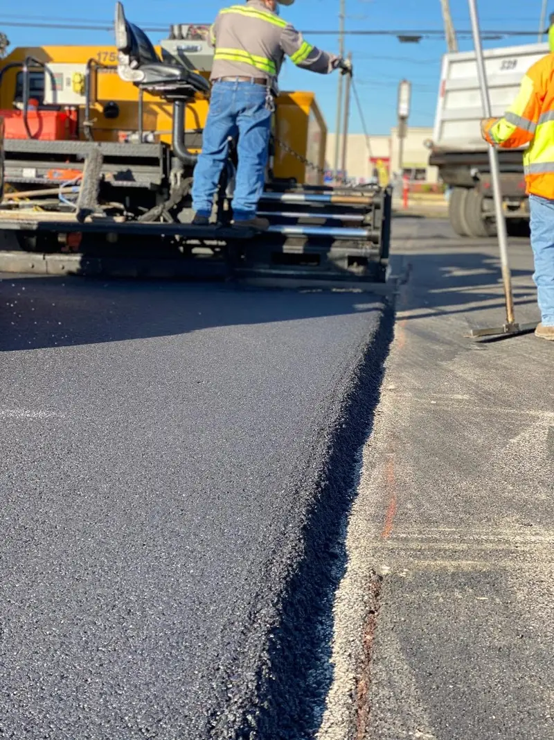 Paving crew running an asphalt paver along a commercial lot, laying a new lift beside the old surface