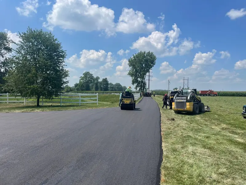 Roller and skid steer finishing a new driveway past a white fence and open farmland