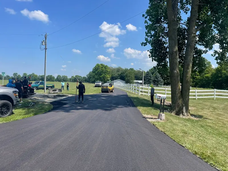 Crew finishing a long new driveway beside a white board fence, with the roller working toward the far end