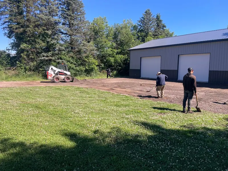 Skid steer and crew grading a crushed-stone base in front of a pole barn before paving