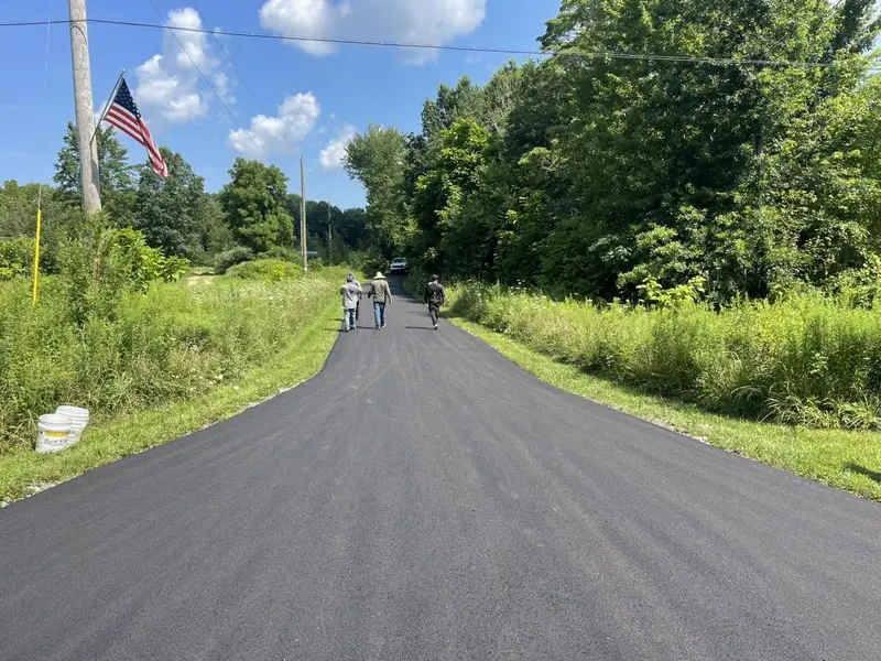 Newly paved lane running between tall summer brush and trees, with the crew walking the finished surface